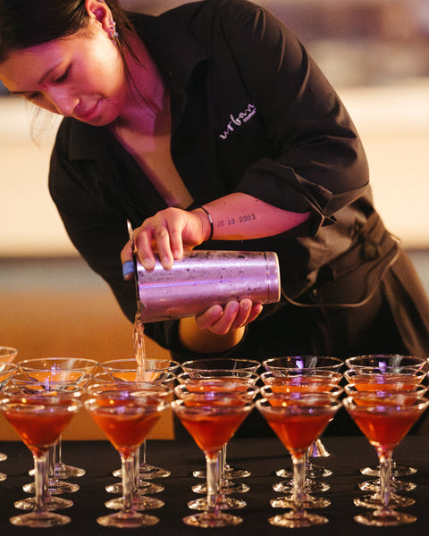 Cocktail server pouring drinks into small glasses at a bar.